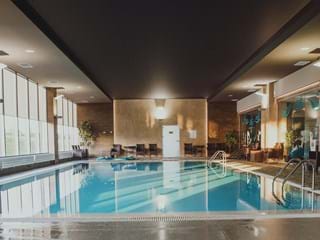 A swimming pool area with soft, natural colours on the floor and walls. Black acoustic panels are installed in the ceiling. To the left there are big windows bringing natural light into the room. Dark brown rattan chairs are placed along the walls and green plants are seen in the corners of the room.