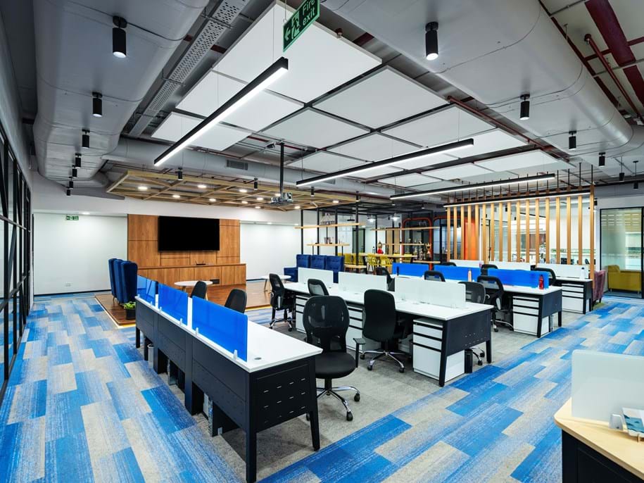 An empty modern office space with blue carpeting. The area is well-lit with overhead lights and features a combination of cubicles with blue partitions and black office chairs. To the left, there is a wooden panel wall with a large screen, and in the background, there are wooden pillars. Visible are also air ducts and free-hanging square acoustic panels in white and grey forming a pattern in the ceiling.