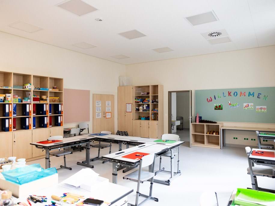 Classroom with birch cabinets and bookshelves along the walls and white desks and chairs in the centre. Acoustic wall panels in pink and turquoise are installed on two of the walls and acoustic panels in the ceiling.