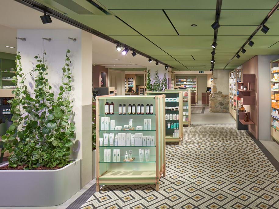 A modern store interior with pale green walls and ceiling, displaying skincare and wellness products on shelves. Potted plants are used as decor. The floor has a geometric patterned carpet. Track lighting illuminates the space.