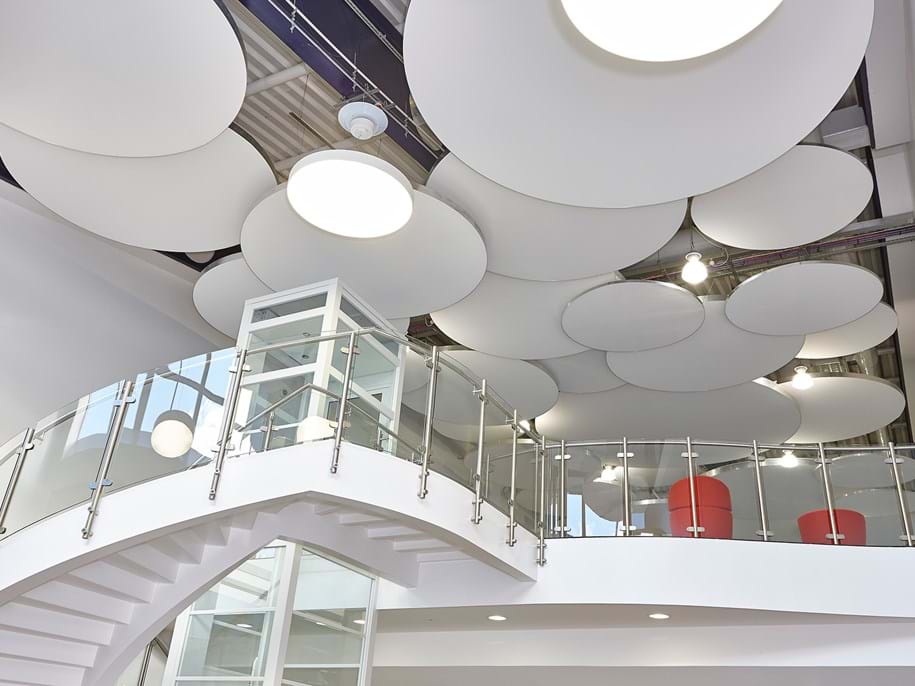 Modern interior with a curved white staircase leading to an upper floor. The ceiling features large, overlapping circular panels and round lights. A glass railing lines the staircase, and red chairs are visible on the upper level.