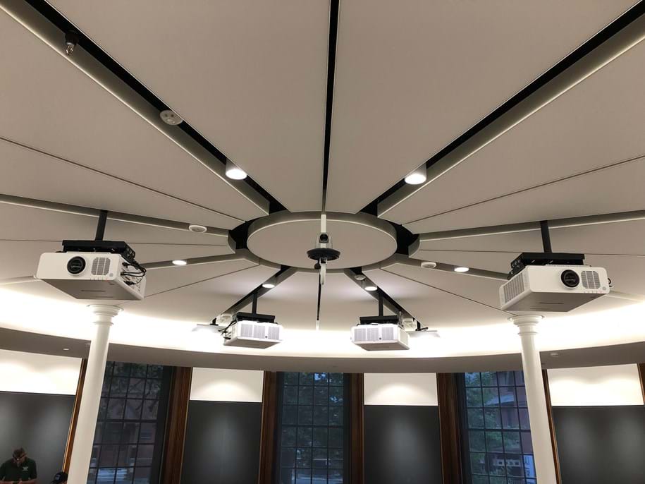 Ceiling view of a room with a central circular structure, consisting of white acoustic panels and multiple video projectors radiating from the center. The room has large windows and modern lighting fixtures.
