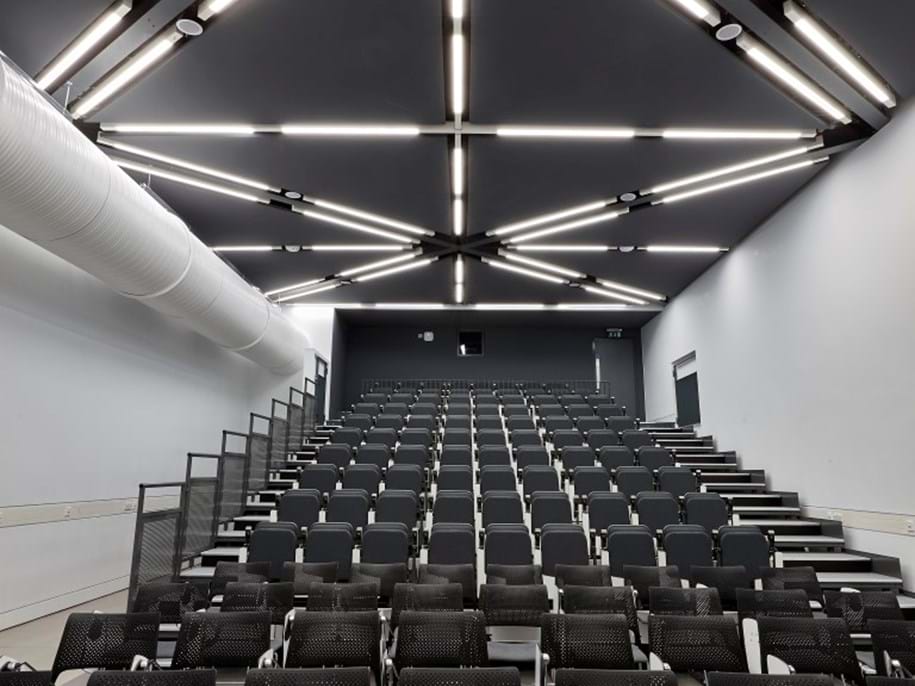 An empty, modern lecture hall with rows of black and grey chairs facing a screen. The ceiling features geometric patterns with bright fluorescent lights integrated in an acoustic ceiling. The walls are plain gray, and there is an air duct on the left side.