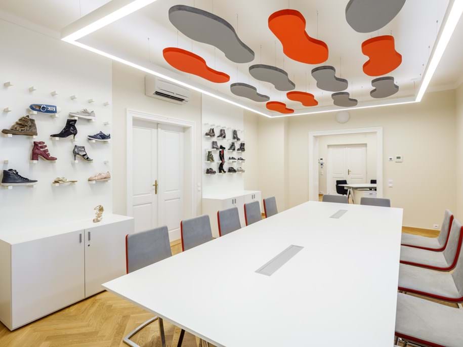 A modern meeting room with a white rectangular table surrounded by red and grey chairs. The room features a unique acoustic ceiling design with grey and orange shoe-like shapes, bright LED lighting, and a display of various shoe models on wall-mounted shelves. There is a door on the far end, leading to another room.
