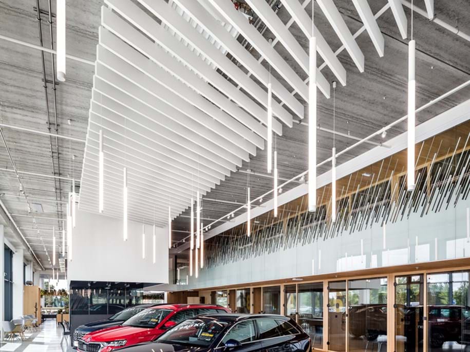 Interior of a modern car showroom with high ceilings featuring an array of white accoustic baffles. Three cars are on display, one in the foreground and two slightly behind it to the left. The floor is polished concrete, and large windows allow natural light to fill the space. In the centre, there is a red rectangular seating area with two round black stools. The showroom appears spacious and designed to highlight the vehicles for sale.