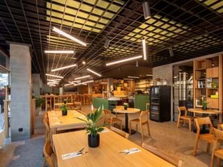 Moderní interiér restaurace s dřevěnými stoly a židlemi, minimalistickým dekorem a rostlinami. Na stropě s mřížkovým vzorem visí jasná svítidla. V pozadí je bar a police s různými předměty.