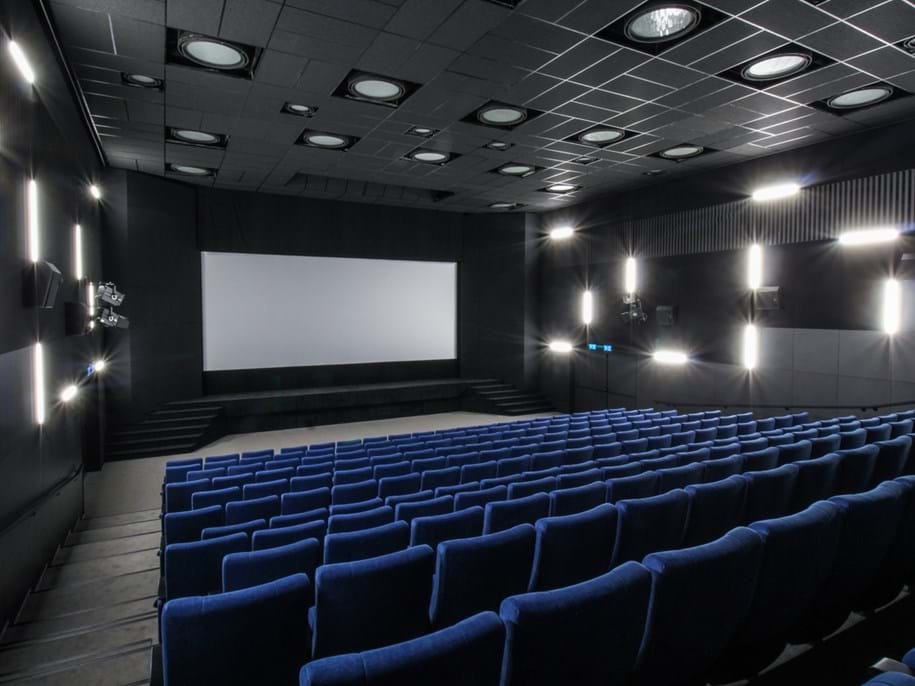 Moderní kino s řadami modrých sedadel obrácených k velkému prázdnému plátnu. Stěny a strop jsou tmavé, vybavené kruhovými světly a reproduktory. Místnost je prázdná, což vytváří tichou a prostornou atmosféru.