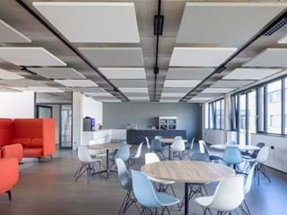 Modern office break room with large windows, white walls, and grey flooring. The space includes a variety of seating options such as round tables with blue and white chairs and red armchairs. The ceiling features square grey and white acoustic panels and there are black light fixtures above. The room is well-lit with natural light and in the background a kitchen area is visible.