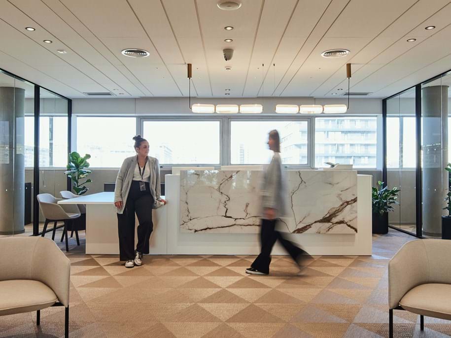 Two people in an office lounge area with modern decor. One person is walking past a marble counter; the other stands nearby. The room has large windows, indoor plants, and a geometric-patterned carpet. Soft lighting creates a calm atmosphere.