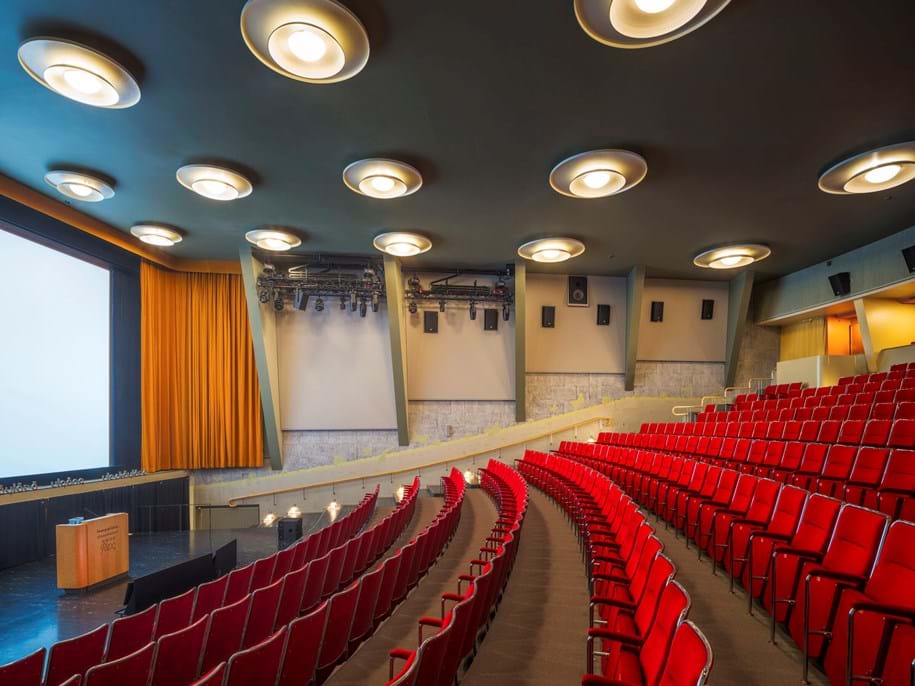 Panneaux muraux acoustiques en tissu extensible dans un cinéma classique avec des rangées de chaises en tissu rouge et un plafond bleu pétrole avec des lampes rondes.