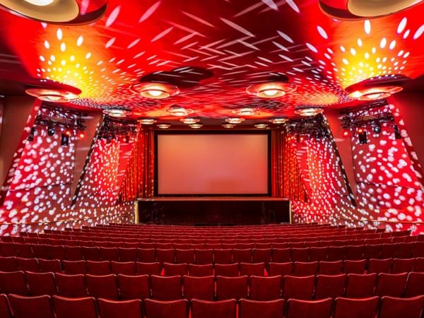 Klassisen elokuvateatterin akustiset joustokankaiset seinäpaneelit, punaiset kangastuolit rivillä suurelle valkokankaalle päin. Katto ja seinät on koristeltu punavalkoisilla valon kuvioilla, jotka luovat eloisan ja dynaamisen tunnelman.