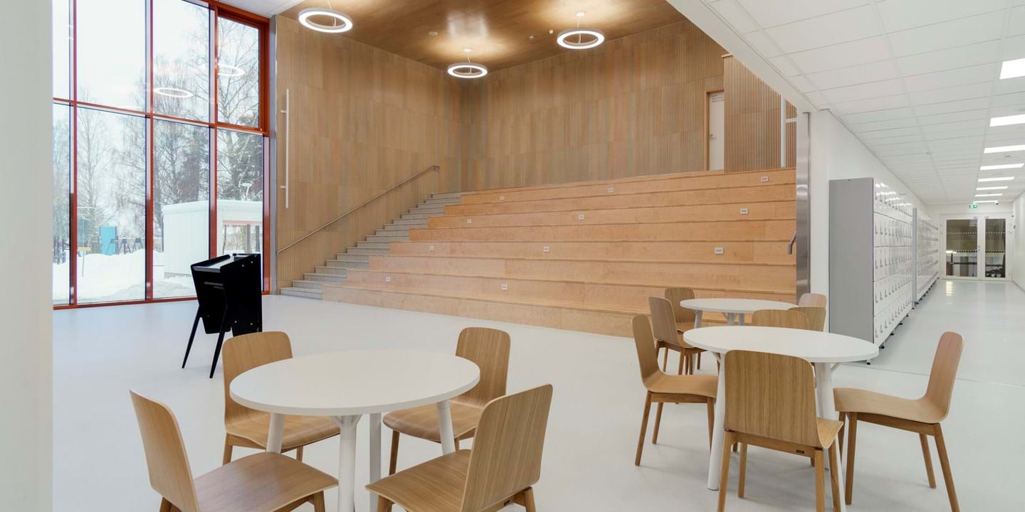 Hämeenlinnan yhteiskoulun uudisrakennuksen aula. Etualalla tuoli- ja pöytäryhmiä, taustalla puiset jättiläisen portaat. Alakatot Ecophonin valkoisia CarbonLow-akustiikkalevyjä.