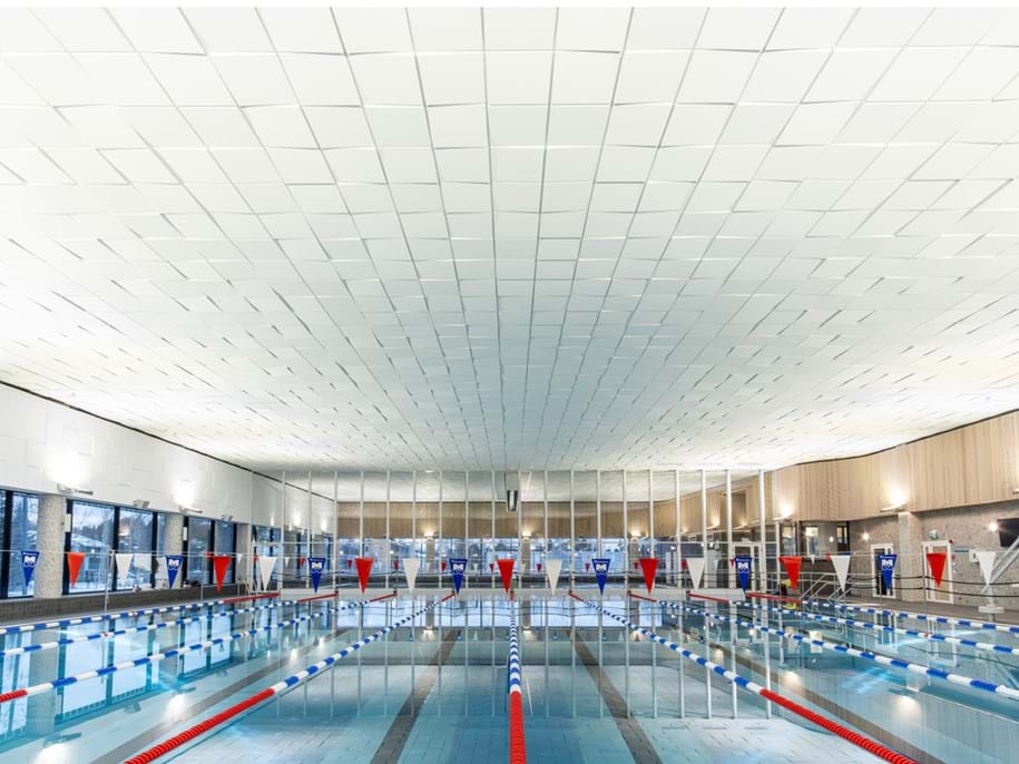 Empty indoor swimming pool with lanes marked by red and blue ropes. Large windows line the sides, and flags are strung above. The ceiling is patterned with square acoustic panels creating an multi-dimensional effect. Bright, even lighting fills the space.