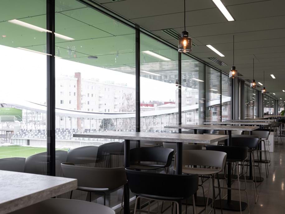 Interior view of a modern cafeteria with a long row of tables and chairs facing large windows that offer a view of an outdoor sports stadium. The space is well-lit with hanging industrial-style light fixtures, and the design features a minimalist aesthetic with a green floor and a light grey acoustic ceiling.