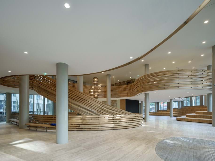 Spacious modern interior with a sweeping wooden spiral staircase, neutral-toned flooring, and large windows. Circular light fixtures hang from the ceiling, and there is seating integrated into the design.