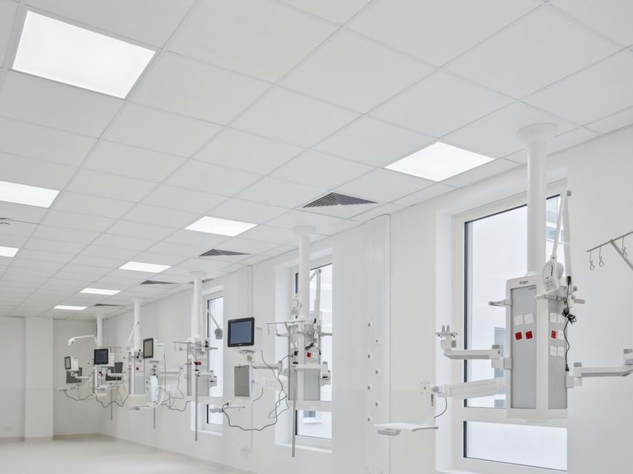 A modern hospital room with multiple medical devices mounted on the ceiling, including monitors and IV stands. The room is brightly lit with natural light from large windows and artificial light from the ceiling panels integrated in the acoustic ceiling. There are no people present in the image.