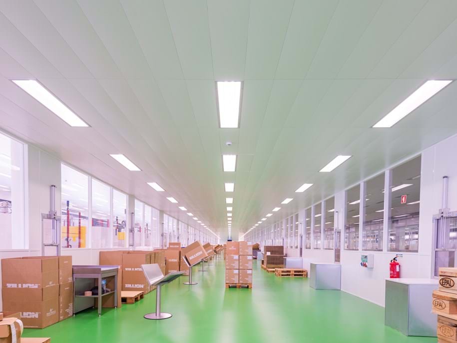 A spacious, well-lit factory interior with a green floor, white walls, and a white metal ceiling featuring bright fluorescent lights. Cardboard boxes and pallets are lined up neatly on both sides of a central aisle.