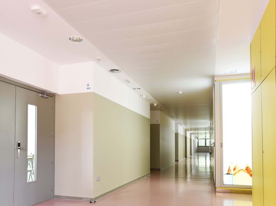 A brightly lit, empty corridor with beige and yellow walls, featuring a closed door with a window on the left, and a large window at the far end. The floor is shiny, reflecting the light from the windows.