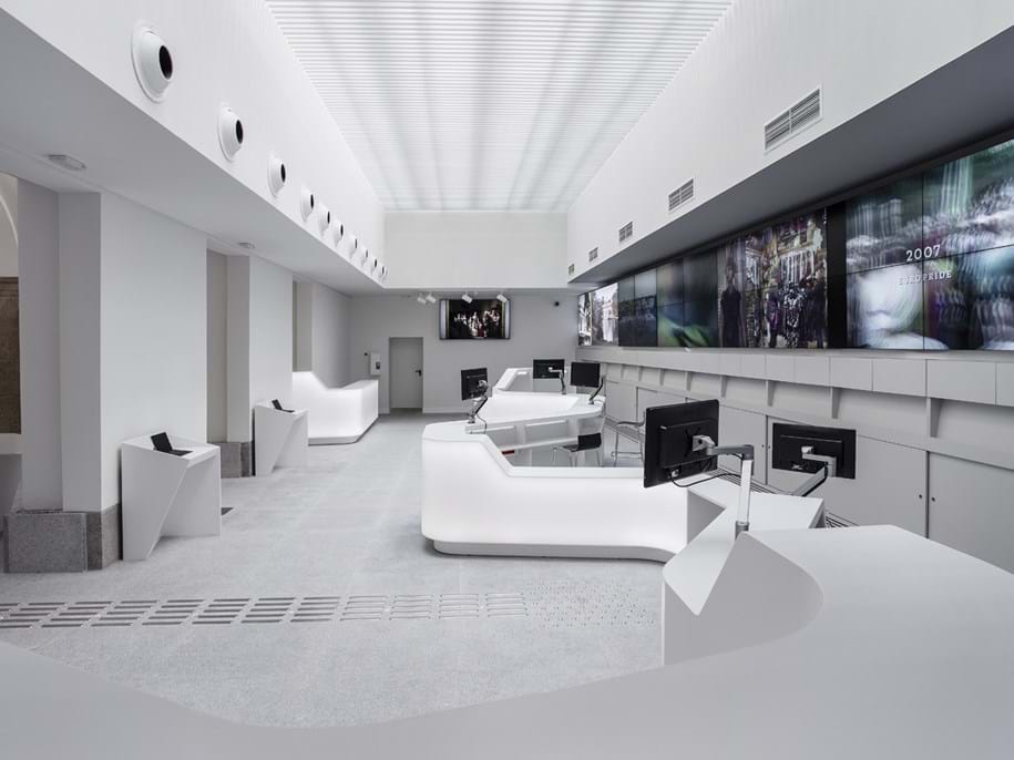 A modern, minimalist room with sleek white desks and multiple flat-screen monitors on the walls. The space is well-lit, featuring a high ceiling with a grid pattern and vent-like details, creating a futuristic atmosphere.