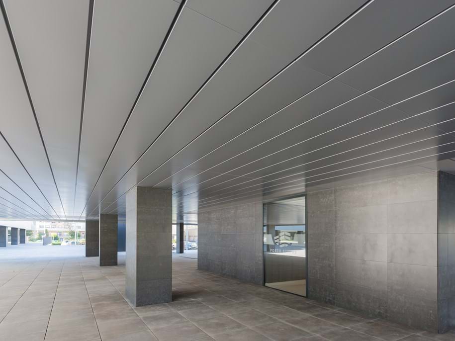 A modern building exterior features large stone columns and geometric lines, creating a sleek, minimalist look. The ceiling and walls are clad in dark, polished panels, and a window reflects light from the open area outside.
