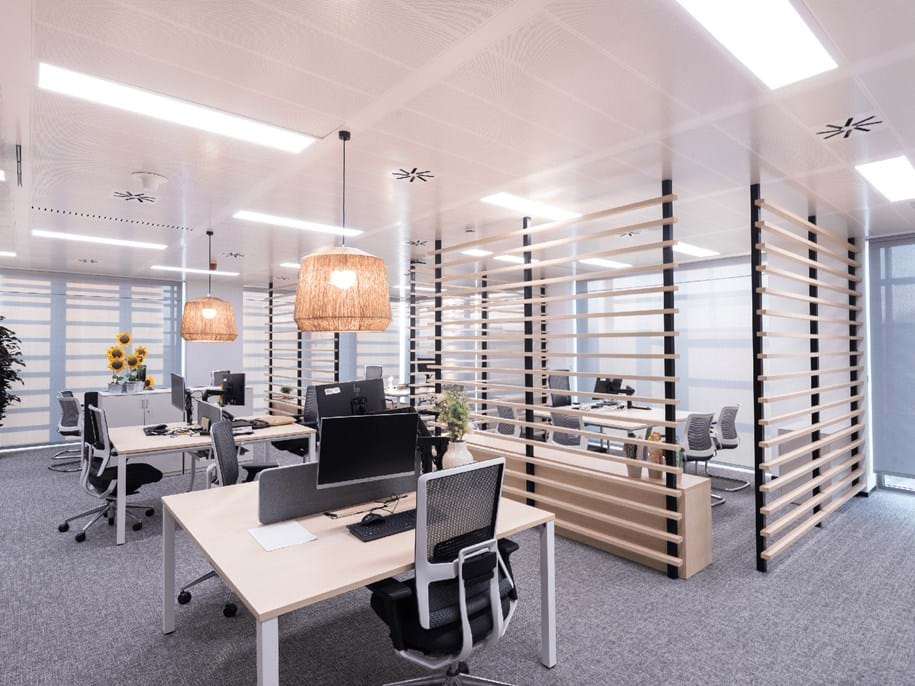 A modern office space with open workstations featuring light wooden desks and ergonomic chairs. The area has partitions with wooden slats, overhead lighting, and large windows with roller blinds, creating a bright and airy atmosphere.