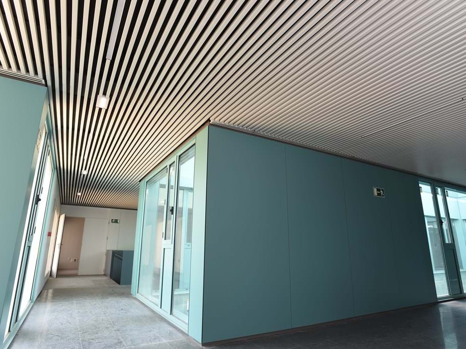 Modern corridor with turquoise walls and a slatted metal ceiling featuring recessed lighting. Glass partitions separate sections, and emergency exit signs are visible. The floor is light gray, reflecting natural and artificial light.