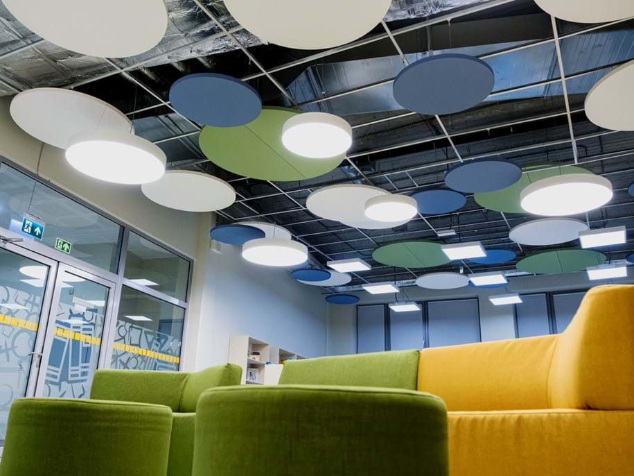 School interior with modern design featuring colourful circular acoustic panels on the ceiling, large green and yellow sofas in the foreground, and glass doors with visible emergency exit signs in the background.