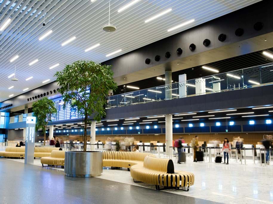 Modern airport terminal interior with high ceilings and bright lighting. Features a large tree in a pot, curved wooden benches, and people standing in line at check-in counters. An information board is visible in the background.