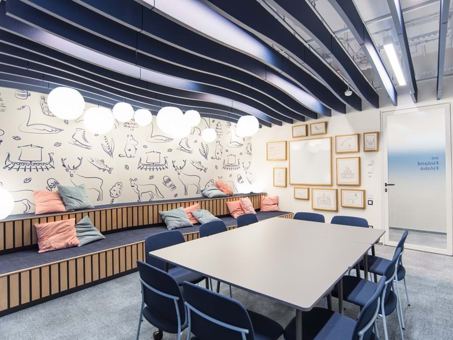 A modern meeting room with a large table surrounded by blue chairs. The room has a wall with playful animal and nature drawings, framed sketches, and tiered bench seating with colourful pillows. Round ceiling lights add to the ambiance.