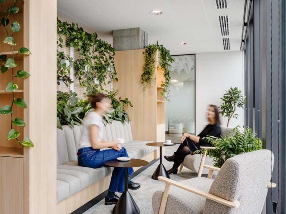 A modern office lounge with two people sitting and talking. The space features wood paneling, grey cushioned seating, small round tables, and abundant green plants. A large window provides natural light.