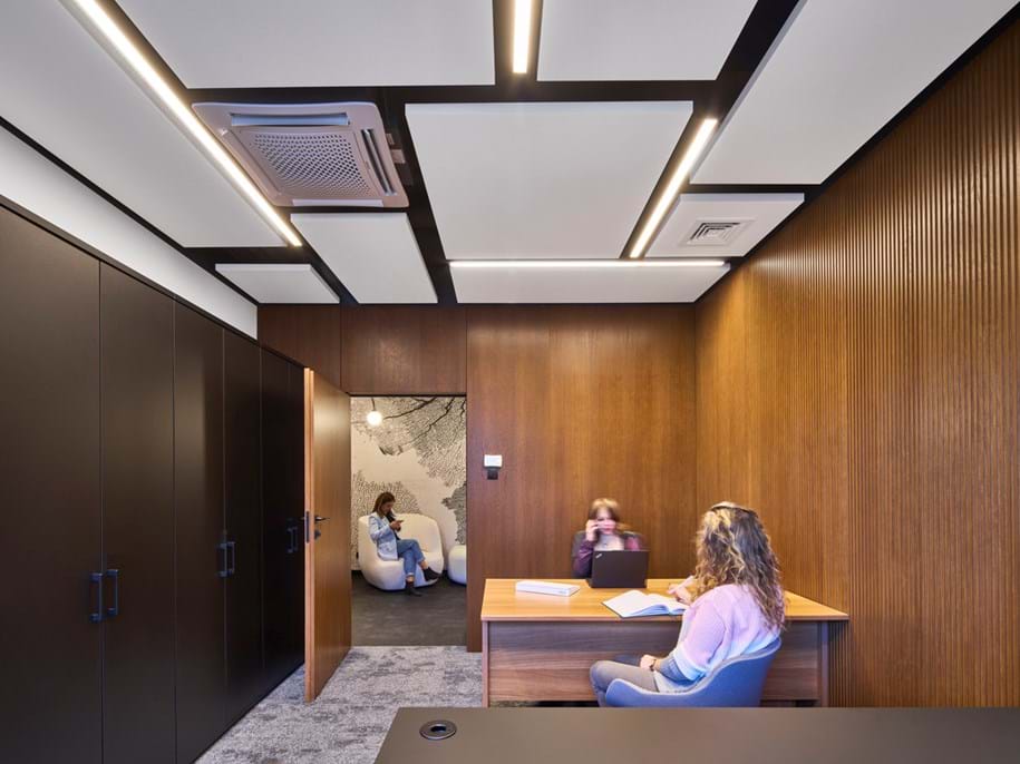 A modern office with wooden walls and ceiling panels. Two people sit at a desk with open books and a laptop. A third person sits on chairs in the adjoining room, visible through an open door. Ceiling lights illuminate the space.