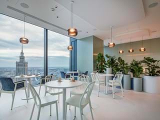 Modern office cafe with white tables and chairs, surrounded by lush green plants in large pots. Floor-to-ceiling windows offer a cityscape view, featuring a prominent tower. Pendant lights hang from the ceiling, enhancing the cozy ambiance.