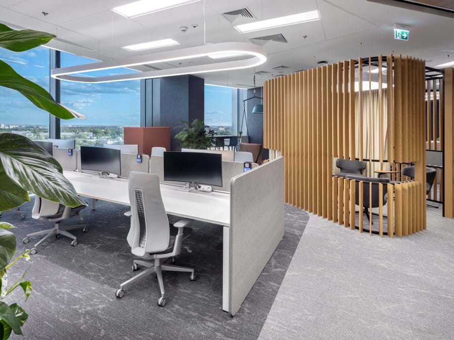 Modern office space with rows of white desks and grey ergonomic chairs. Vertical wooden slats define a seating area. Large windows provide natural light, with a view of greenery outside. Ceiling lights and a plant enhance the rooms ambiance.