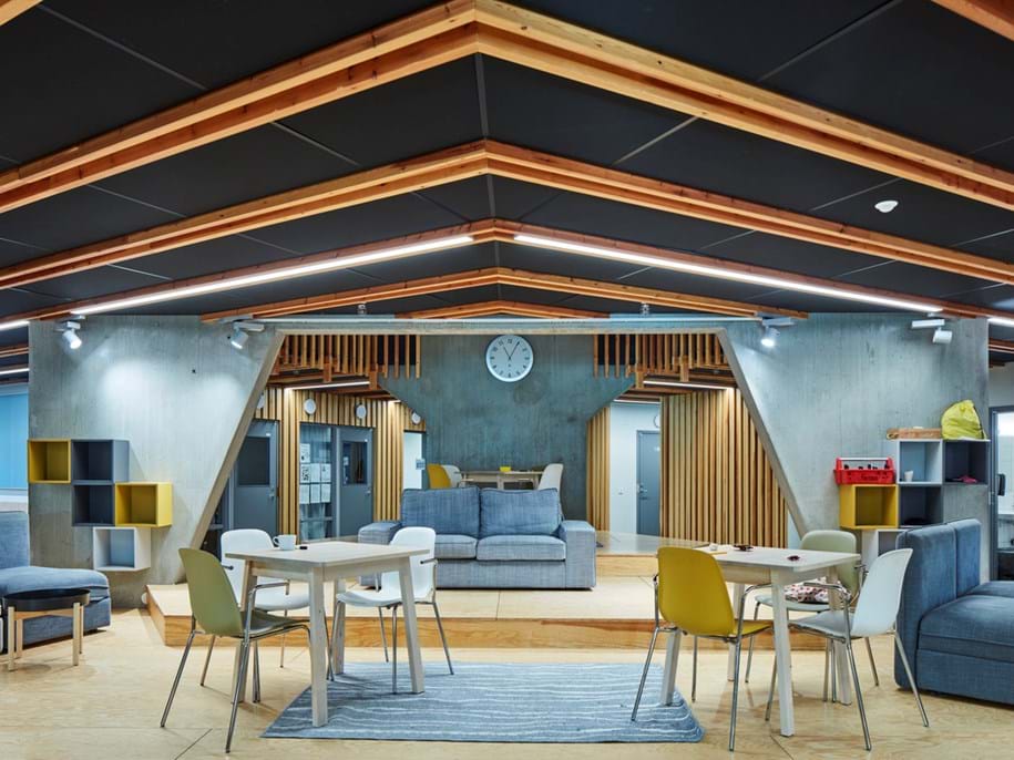 Modern skollokal med geometriska takbjälkar mot ett svart akustiskt tak, en väggklocka och olika sittgrupper. Vita bord, gula och grå stolar, blå soffor och öppna hyllor på väggarna skapar ett inbjudande utrymme för samarbete.