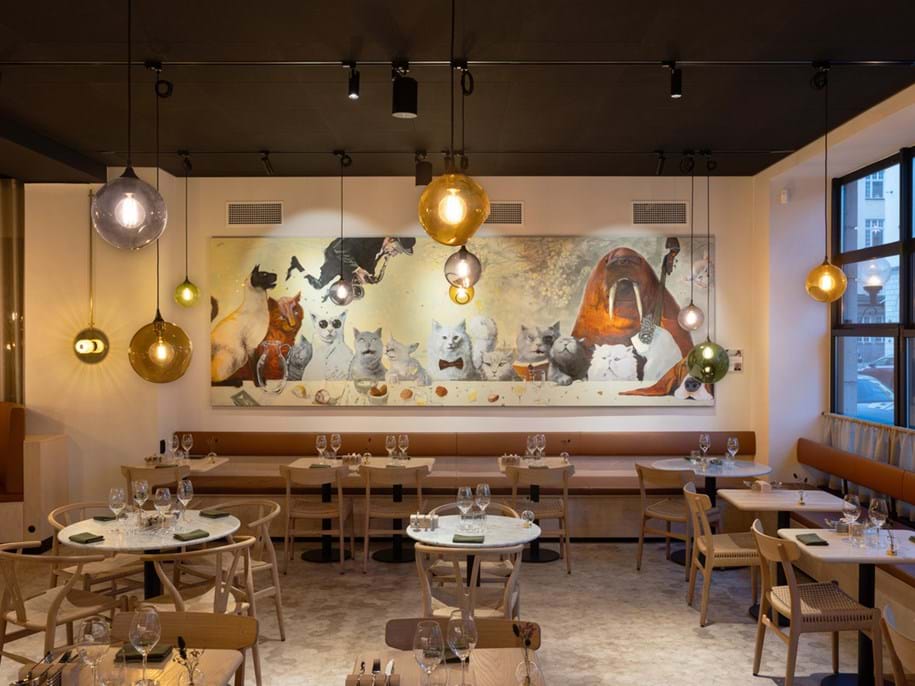Modern restaurant interior with wooden tables and chairs, decorated with various round pendant lights. A mural features cats in whimsical attire. Tables are set with wine glasses and napkins. Large windows show an urban street outside.