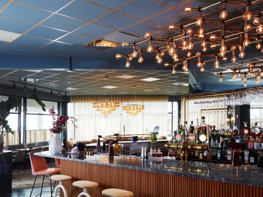 Modern bar interior with a marble countertop, wooden stools, bottles and glasses displayed behind the bar, contemporary lighting fixtures, and large windows with sheer curtains allowing natural light to enter.