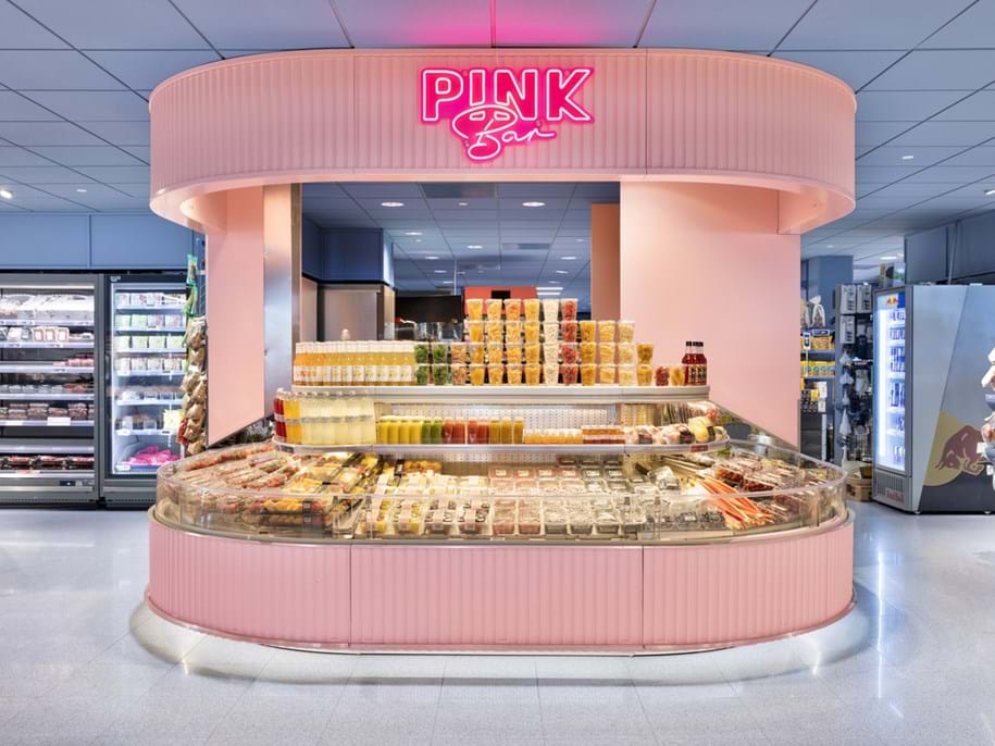 Kirkkaasti valaistu ruokakauppaosasto, jossa on vaaleanpunainen Pink Bar -neonkyltti. Pyöreässä kylmätiskissä on erilaisia pakattuja susheja, mehuja ja salaatteja. Taustalla näkyy hyllyjä, joissa on juomia ja välipaloja.
