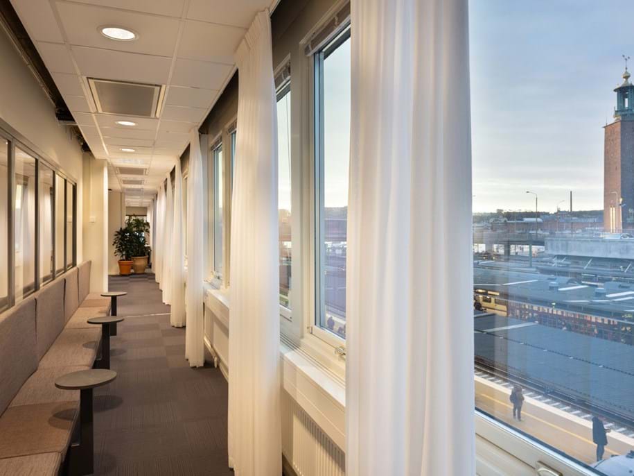 A bright corridor with large windows covered by sheer curtains overlooks a cityscape, featuring a prominent clock tower. The hallway has chairs and small tables along the wall. The sky is clear, indicating daytime.