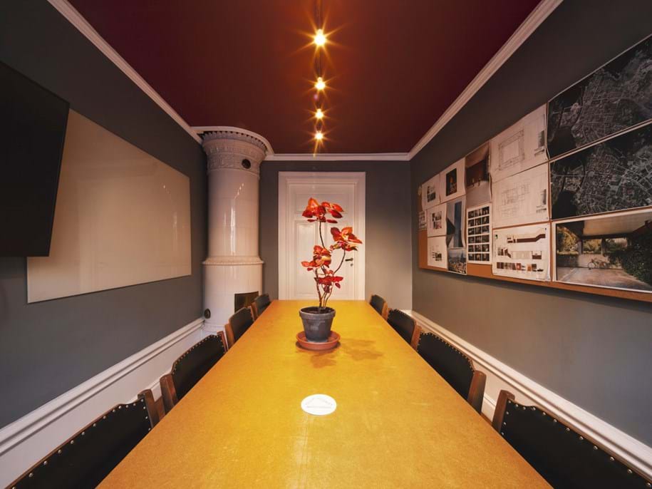 A long wooden conference table with a potted red plant in the centre. The room has dark grey walls adorned with architectural plans, a TV, and warm overhead lighting in the dark red ceiling. A traditional white stove is visible at one end.