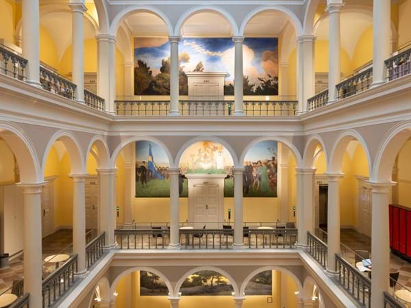 Interior view of a grand, multilevel building with arched balconies and a central atrium. The walls are adorned with colourful murals, and red lockers line the lower level. Warm lighting enhances the architectural details.