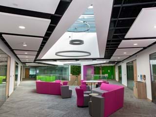 Modern office interior with a skylight, featuring pink and grey seating areas. The space is lined with glass walls and vibrant green accents, creating an open and airy atmosphere. Circular light fixtures hang from the ceiling.
