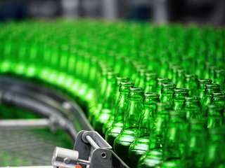 Rows of empty green glass bottles move along a conveyor belt in a beverage or bottling factory, with the bottles closely aligned and the background slightly blurred.