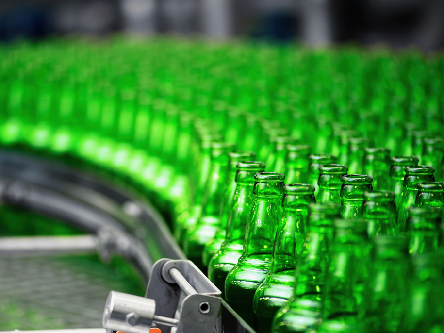Rows of empty green glass bottles move along a conveyor belt in a beverage or bottling factory, with the bottles closely aligned and the background slightly blurred.