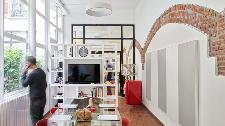 Moderni avotilatoimisto, jossa on suuri lasipöytä ja punaiset tuolit. Valkoisella hyllyllä on musta televisio ja erilaisia sisustusesineitä. Henkilö seisoo korkeiden ikkunoiden ääressä, joista on näkymä ulos. Valkoisia seiniä koristavat tiilikaaret, ja nurkassa on punainen jääkaappi.