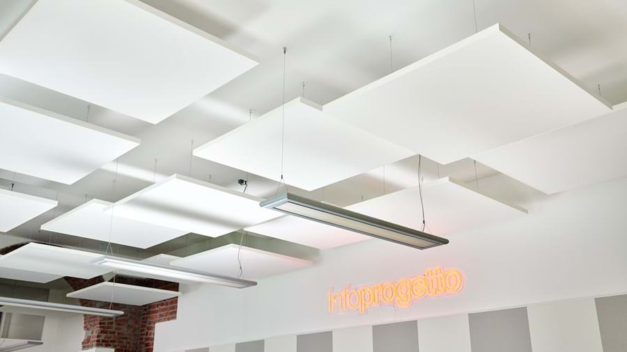 Modernissa tilassa oleva alakatto, jossa on suorakaiteen muotoiset akustiikkalevyt sekä riippuvalaisimia. Seinällä olevassa neonkyltissä lukee oranssilla Infoprogetto. Seinä, jossa on punaisia tiilitehosteita ja valkoisia seinälevyjä.