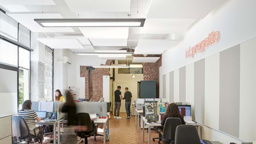 Avoin toimistotila, jossa ihmiset työskentelevät tietokoneilla varustettujen työpöytien ääressä. Huoneessa on valkoiset seinät, tiiliaksentteja ja moderni valaistus. Kaksi ihmistä seisoo ja puhuu taustalla lähellä sinistä ovea.