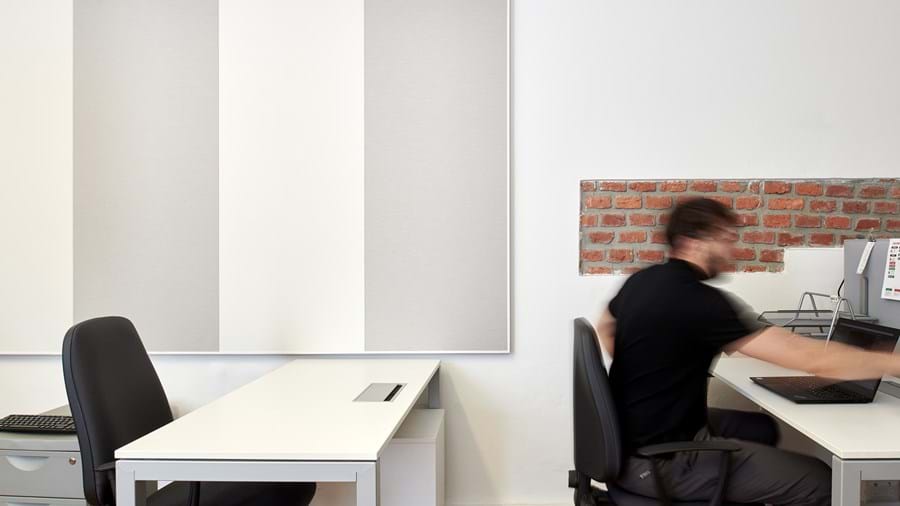 Mustapaitainen henkilö liikkuu nopeasti modernissa toimistotilassa, jossa on valkoisia pöytiä ja tuoleja. Taustalla näkyy tiiltä ja valkoista yhdistävä seinä sekä pystysuoria kangaspaneeleja. Tilanne vaikuttaa dynaamiselta, ja siinä on liikkeen tuntu.