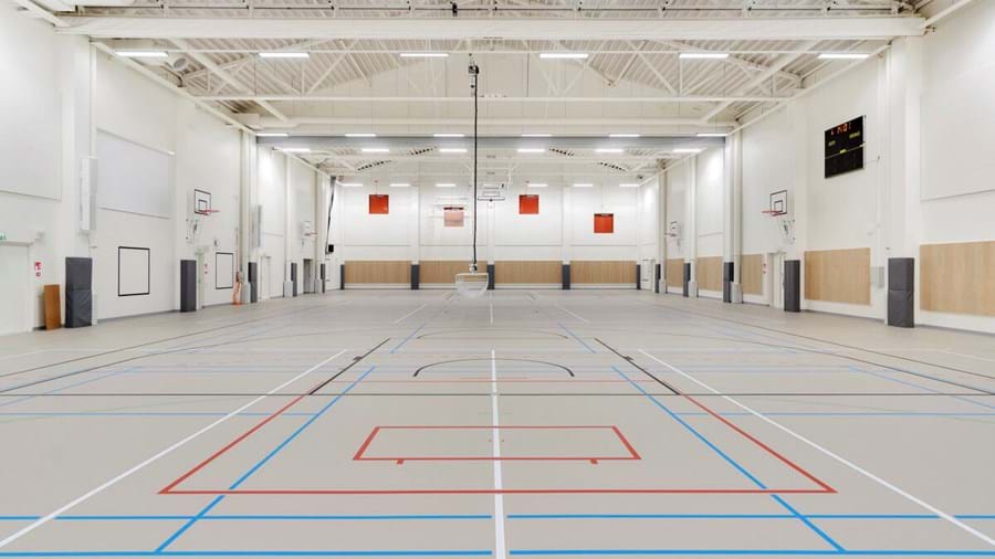 Didelės tuščios gimnazijos salės interjeras su keliomis krepšinio aikštelėmis su raudonomis ir mėlynomis linijomis. Lubos yra aukštos, matomos sijos, o dešinėje pusėje yra prie sienos pritvirtinti lankai ir švieslentė.