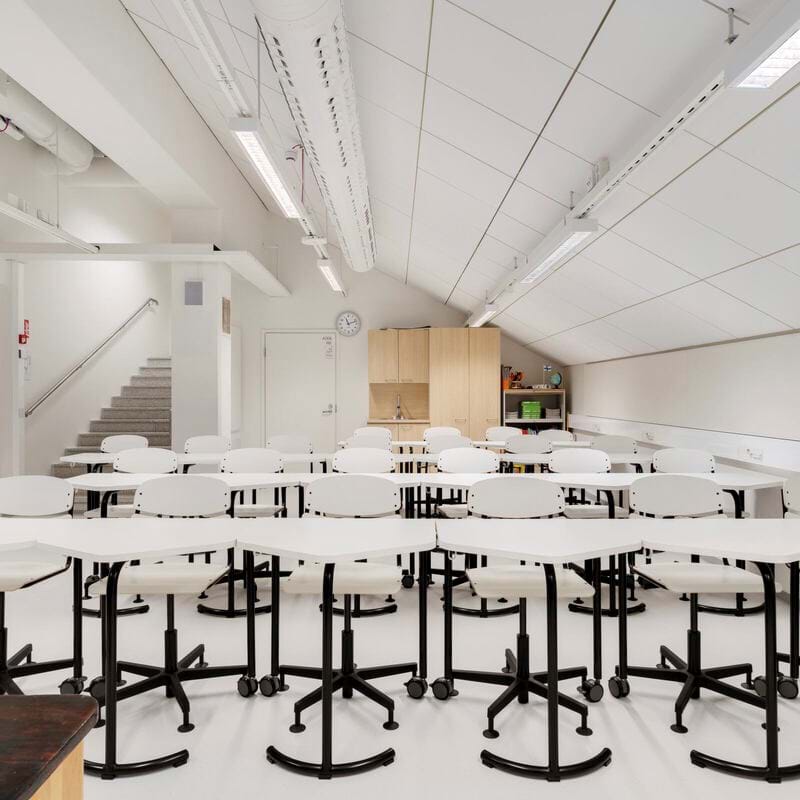 Tuščia klasė su baltomis kėdėmis ir eilėmis sustatytais stalais. Kambaryje yra nuožulnios lubos, fluorescencinės lempos ir sieninis laikrodis. Fone matomi laiptai, vedantys į kitą lygį.