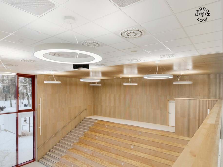 A modern, open indoor space with wooden tiered seating, circular ceiling lights, a red-framed glass wall, and light wood paneling. Snow is visible outside through the windows. Carbon Low is written on the ceiling.